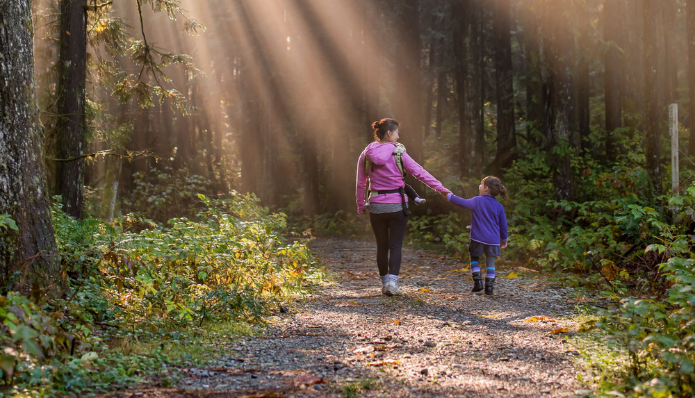 A mother wearing a pink jacket walks with her daughter down a nature trail.